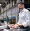 Man in a white shirt working on a laptop in an industrial factory setting