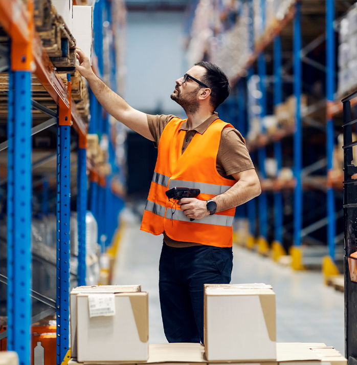Warehouse worker in an orange safety vest using a barcode scanner to manage inventory on high shelves