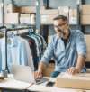 Lächelnder Mann mit Brille arbeitet an einem Laptop in einem Bekleidungs-Webshop, umgeben von Kartons und hängender Kleidung.