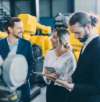 Three colleagues in a discussion, standing in front of yellow industrial machinery, with one person holding a tablet