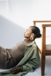 Fashionable woman with sleek hair, wearing large geometric earrings, leaning against a wooden chair in soft natural light.